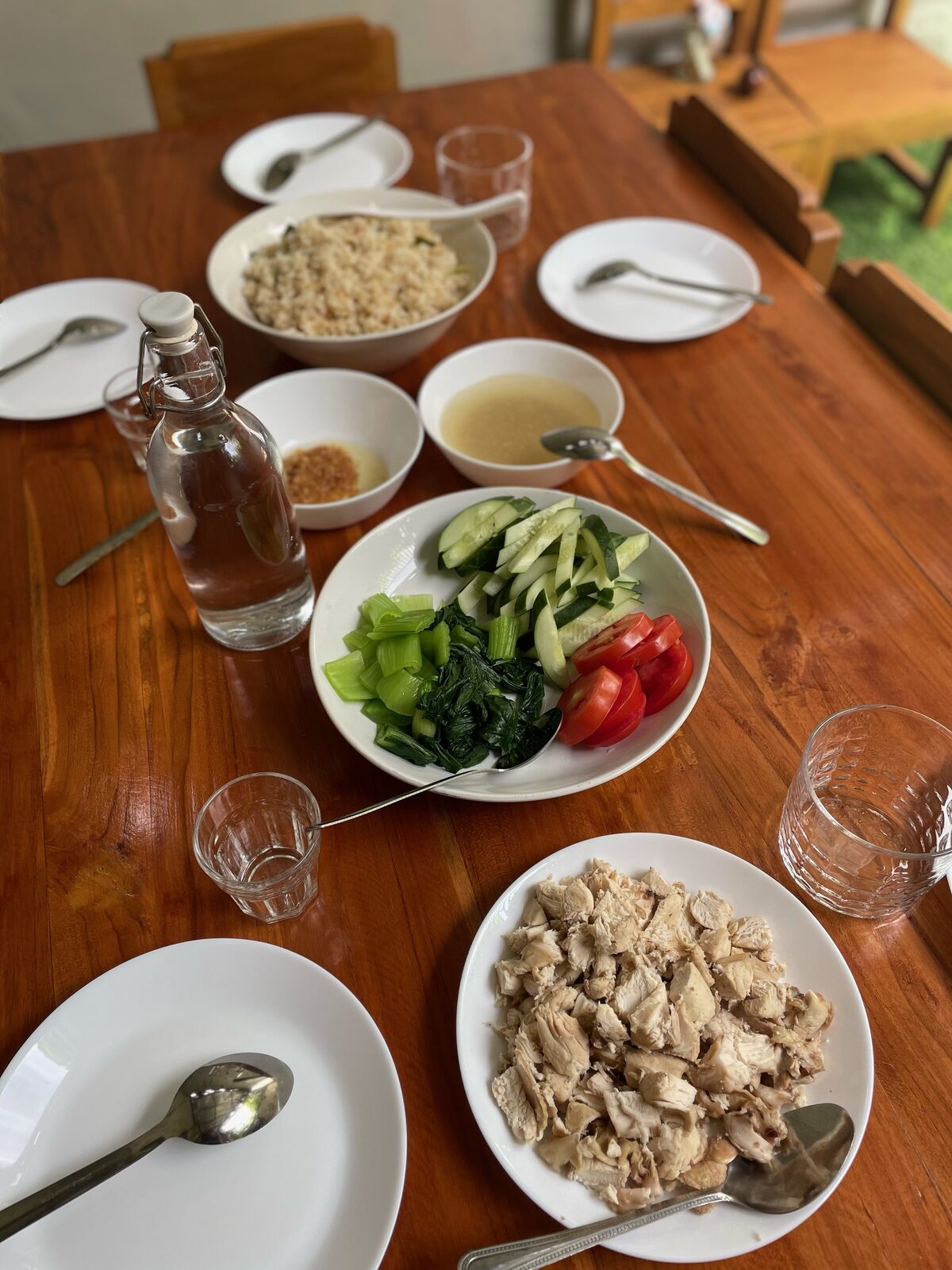 A shared lunch table with rice, clear soup, chicken, bok choy, cucumber and tomato served family-style
