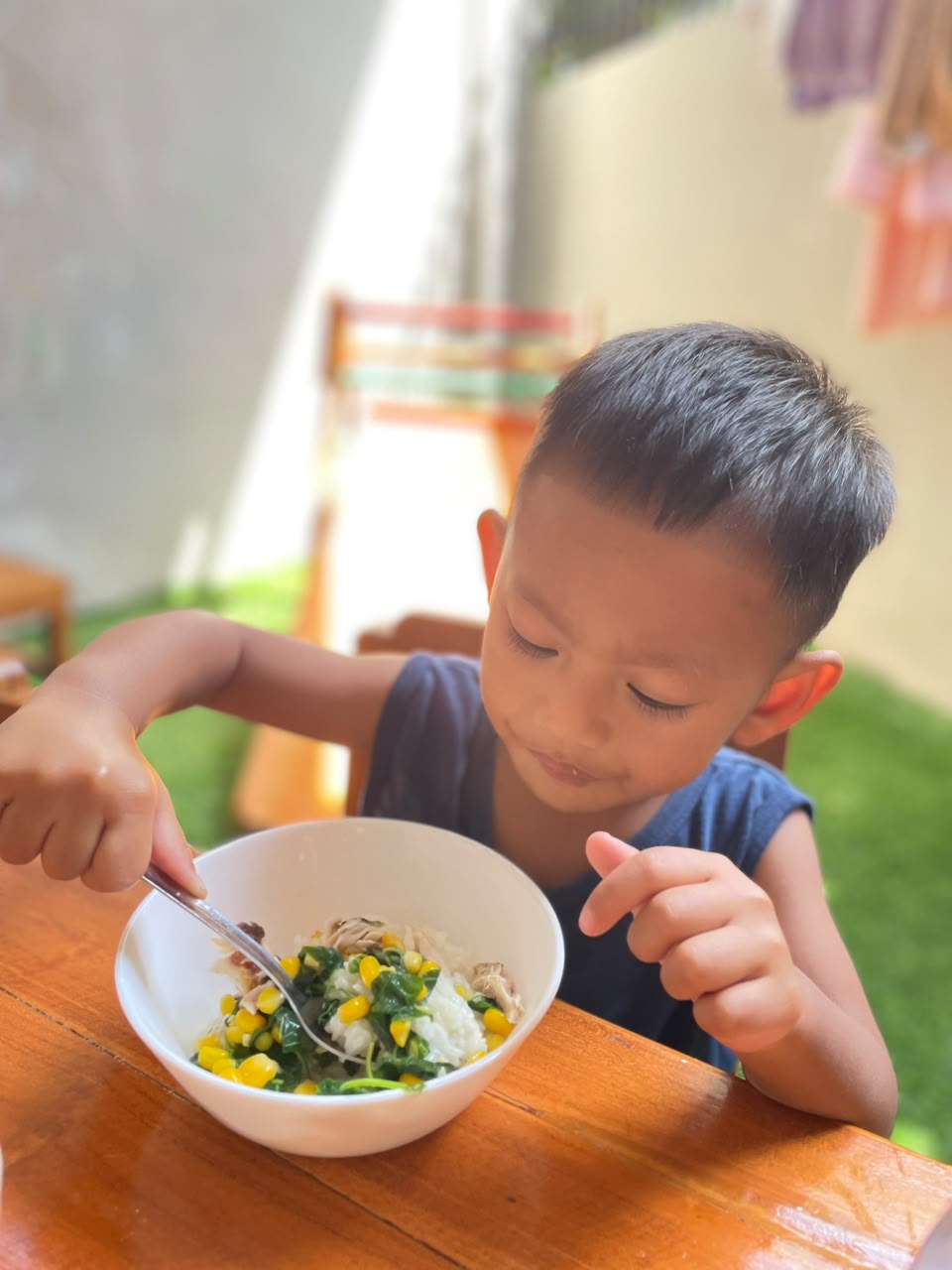 A child at Untuk Anak eating a bowl of rice, spinach and corn at an outdoor table