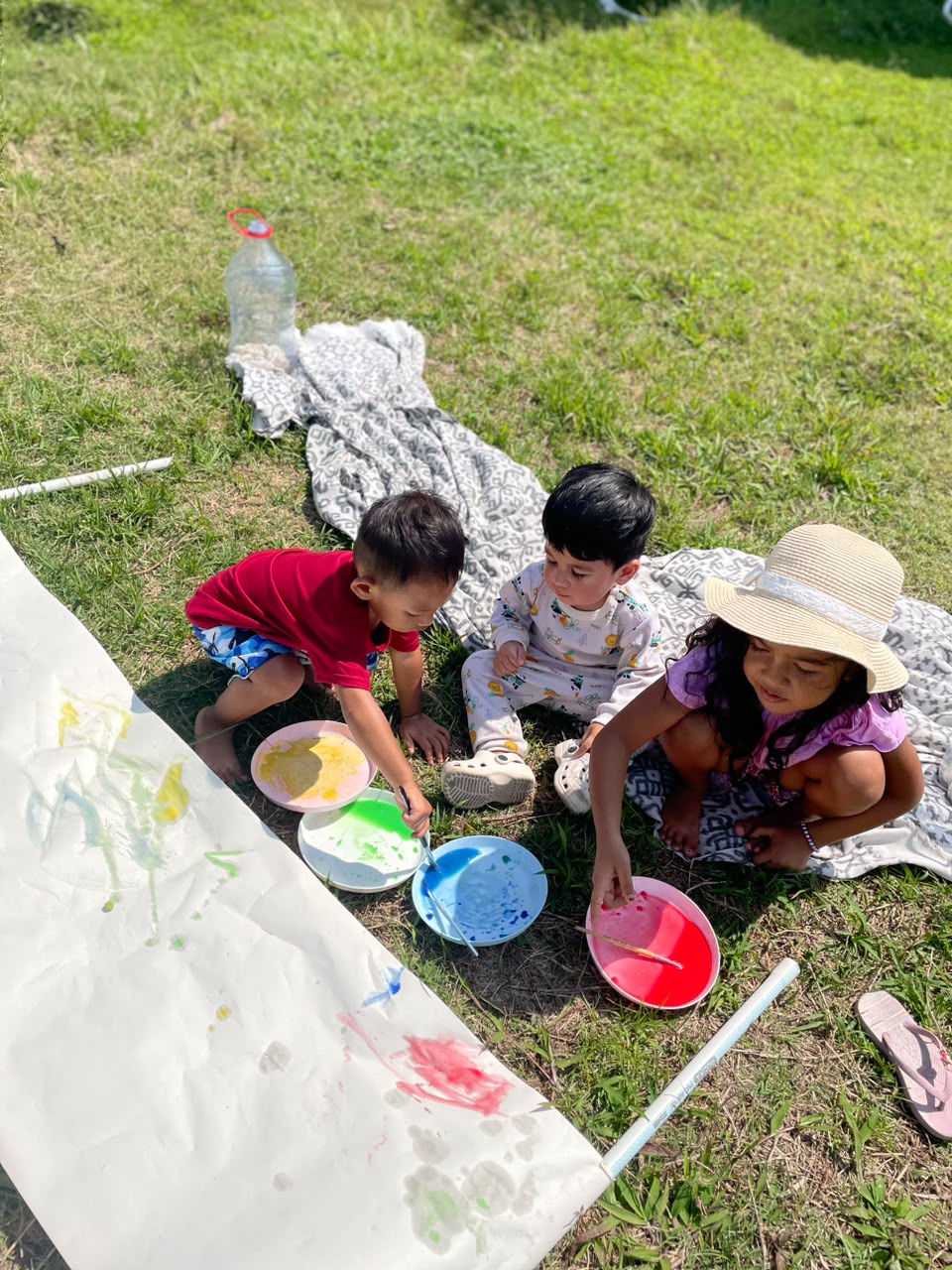 Three children painting with bright colours on a long roll of paper outdoors on the grass