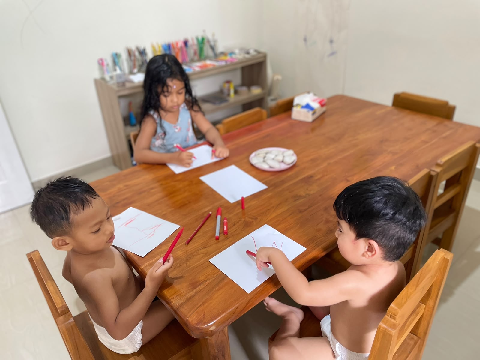 Three children drawing together at a wooden table with coloured pencils