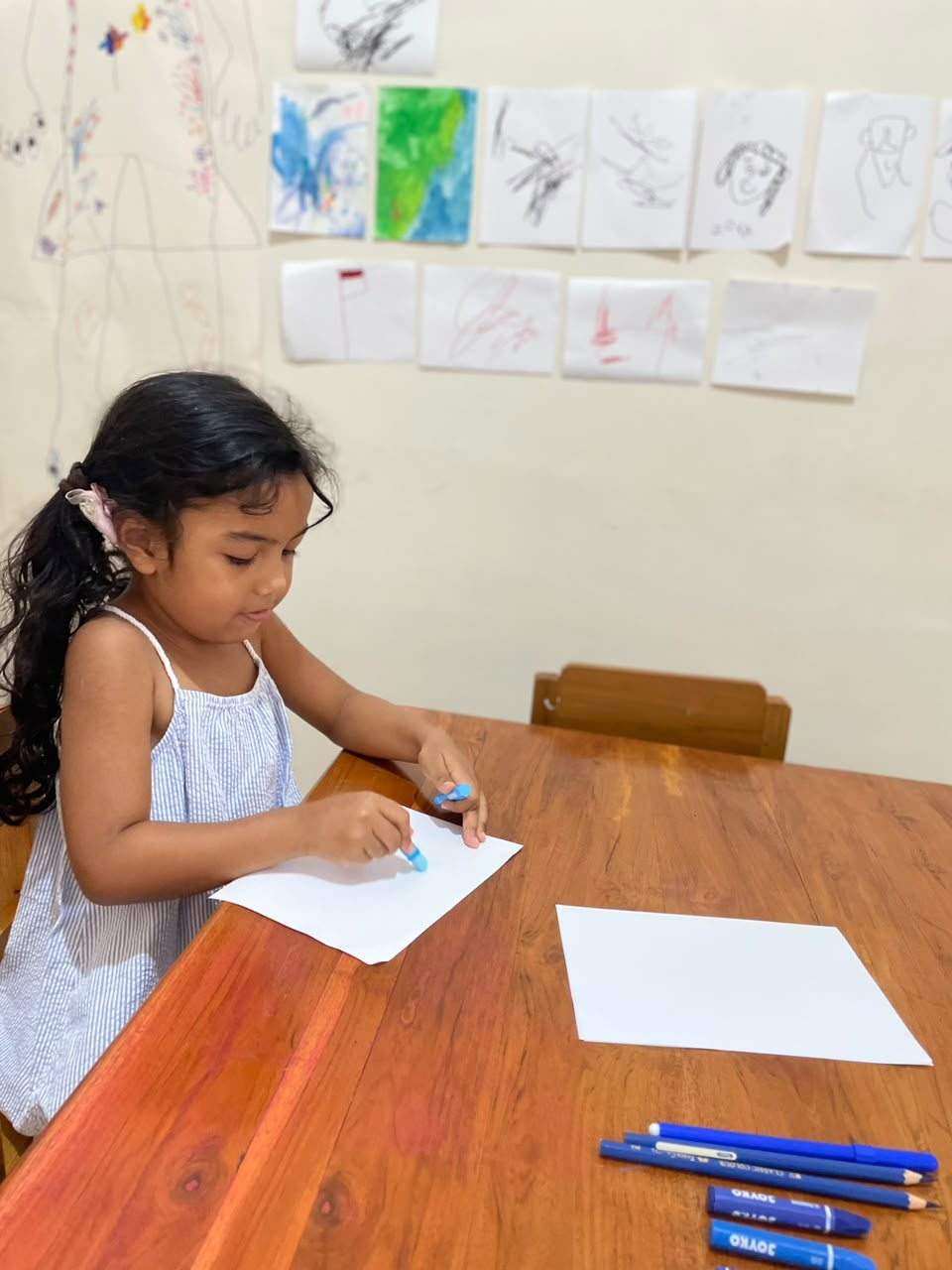 A child drawing with a blue crayon at a wooden table below a wall of children's artwork