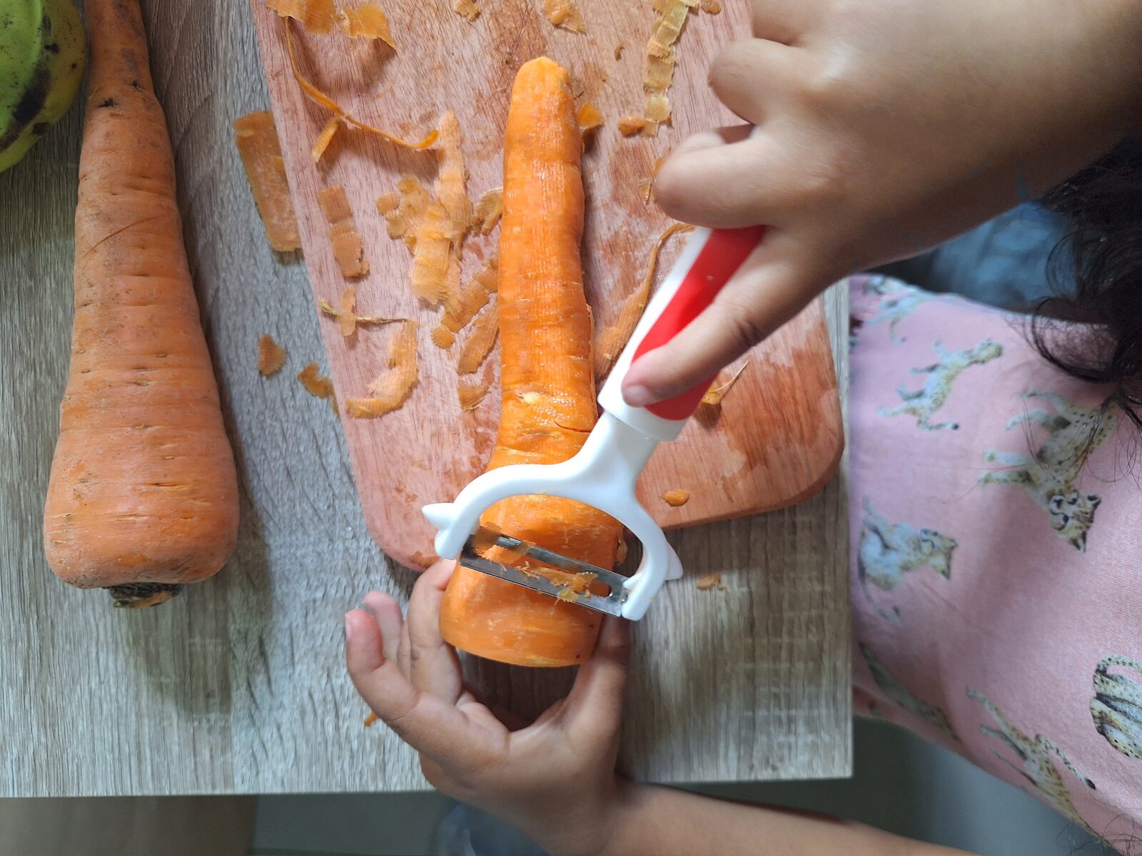 A child peeling a fresh carrot with a vegetable peeler on a wooden board at Untuk Anak