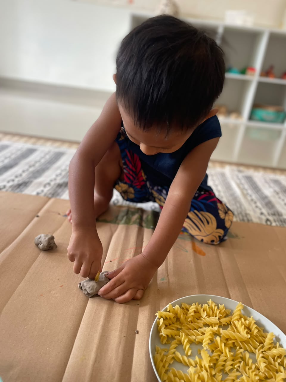 A young child pressing clay onto cardboard beside a bowl of pasta shapes
